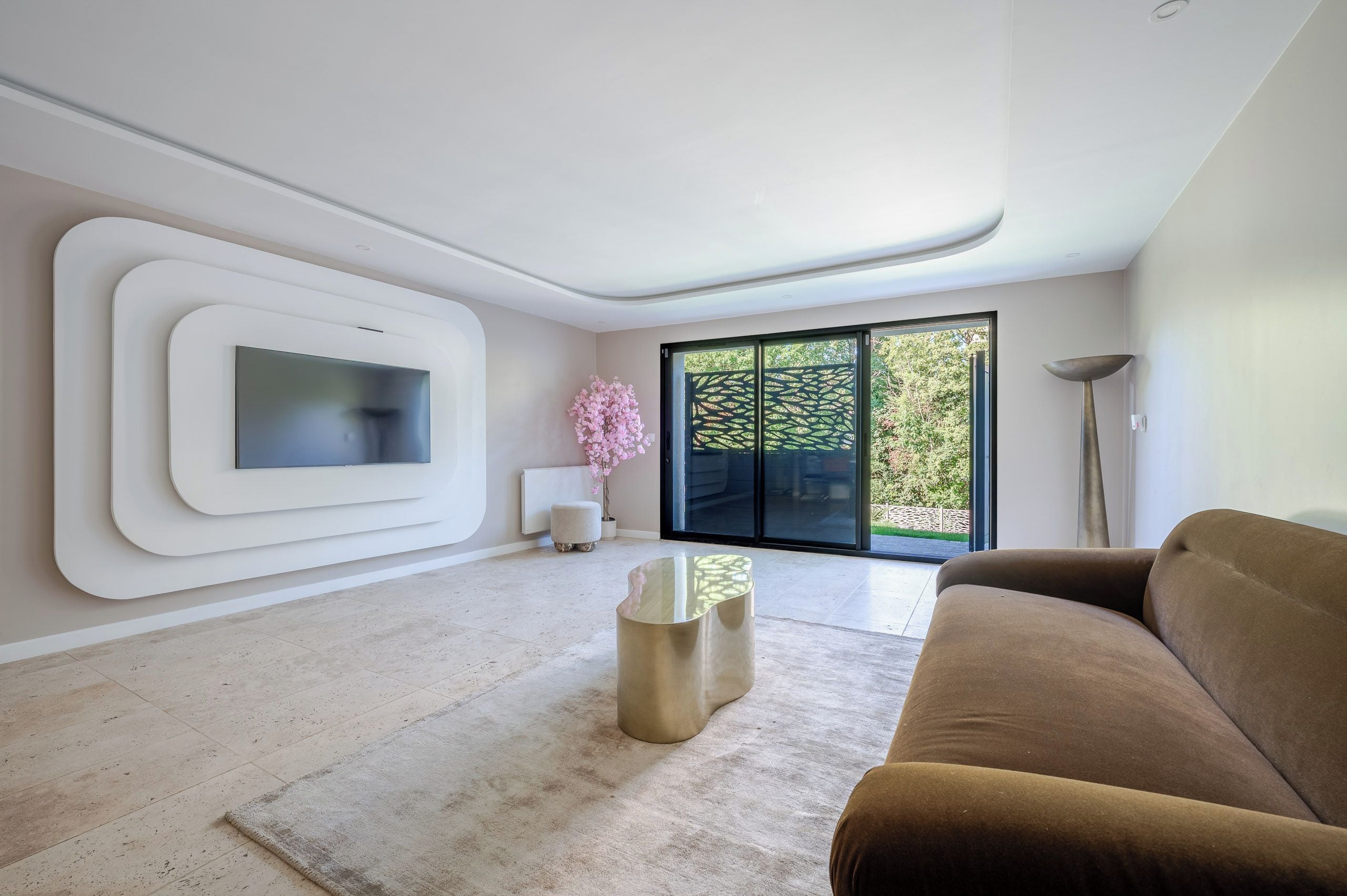 Un salon moderne doté d’un téléviseur à écran plat, d’un canapé marron, d’une table basse en verre et d’une grande porte-fenêtre coulissante donnant sur un jardin.