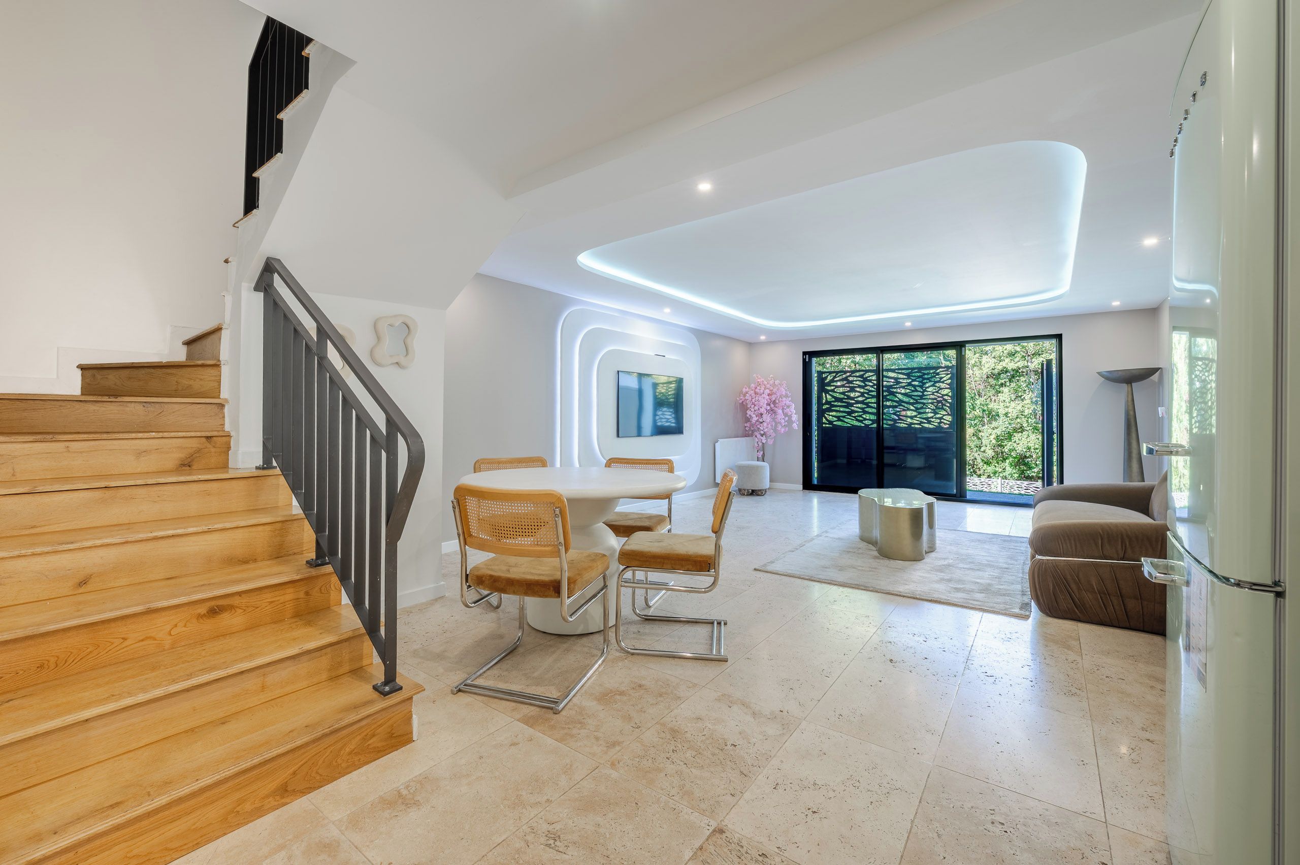 A modern interior with a wooden staircase, a dining area with a round table and chairs, and a large window offering a view of greenery.
