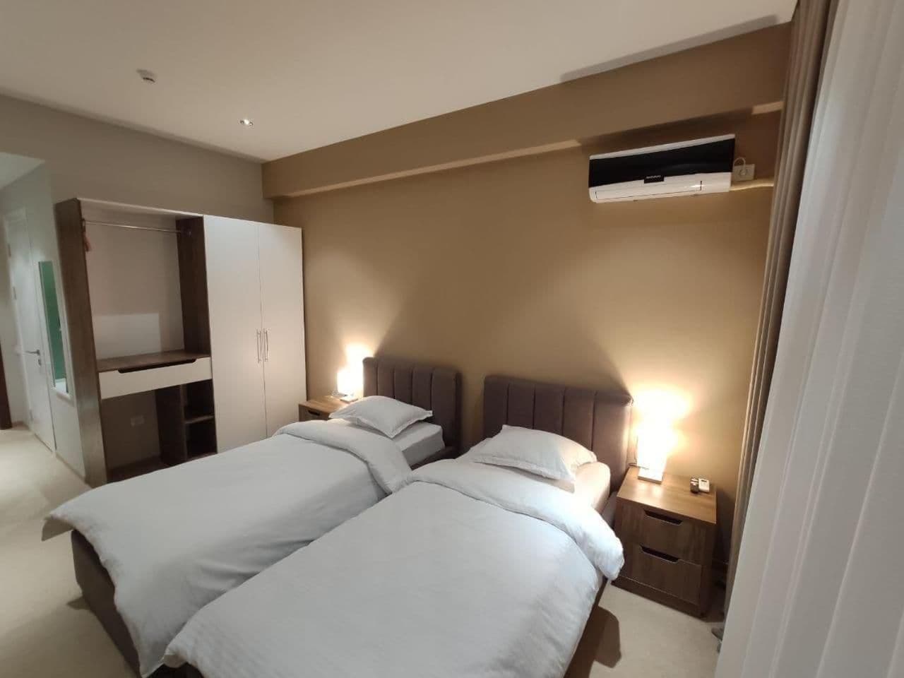 A hotel room with two beds, a wardrobe, bedside tables, and an air conditioning unit.