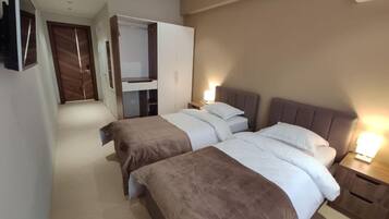 A hotel room with two beds, a wardrobe, a TV, and a bedside table with a lamp.