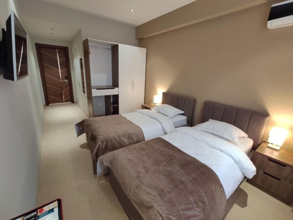 A hotel room with two beds, a wardrobe, a TV, and a bedside table with a lamp. - Pana Hotel (Nukus)