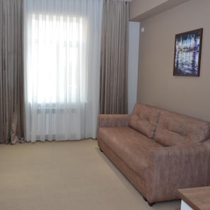 A living room with a beige sofa, a window with sheer curtains, and a painting on the wall. - Pana Hotel (Nukus)