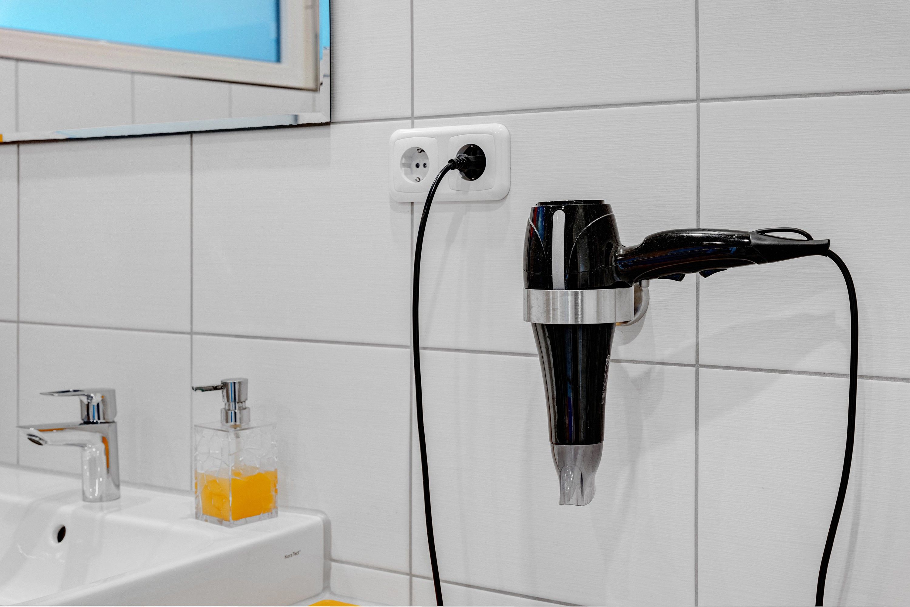Ein Badezimmer mit einem an der Wand befestigten Fön, einem Waschbecken mit Seifenspender und einem Spiegel über dem Waschbecken.