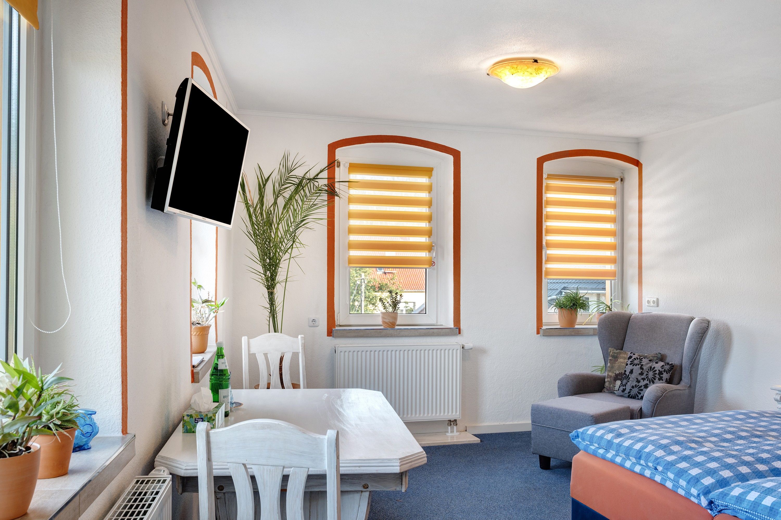 Ein Zimmer mit einem Bett, einem Sessel, einem Tisch, einem an der Wand befestigten Fernseher, zwei Fenstern mit Jalousien und Topfpflanzen.