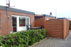 Ein Backsteinhaus mit Holzzaun, einer Terrasse mit Tisch und Stühlen und einer Hecke davor.