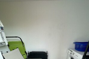 Een wasruimte met een wasmachine, een groene stoel, een blauwe wasmand en een witte plank met een doos waarop 'GIERSCH' staat.