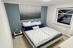 Ein Schlafzimmer mit einem Bett, einem Nachttisch, einer Kommode, einem Fenster und einem Wandbild.