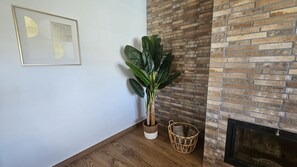 A corner room with a white wall, a framed abstract artwork, a large green plant in a white pot, a wicker basket with items, and a brick fireplace. - Alma del Olivar Holiday Home (La Puebla de Cazalla)