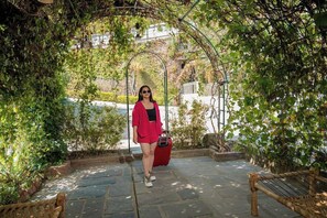 A person standing under a wooden archway in a garden area with stone flooring and a bench.
