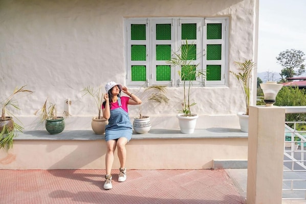 Una persona sentada en un banco, con plantas en macetas, una pared blanca y un ventanal con vidrios verdes.