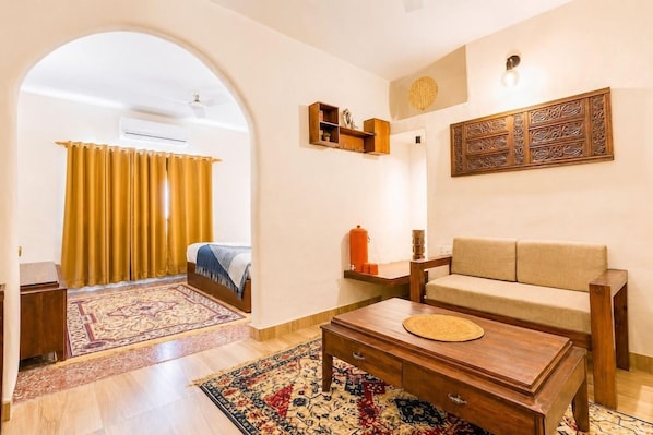 A room with a sofa, a wooden coffee table, a patterned rug, a small shelf with decorative items, and a wall-mounted light fixture.