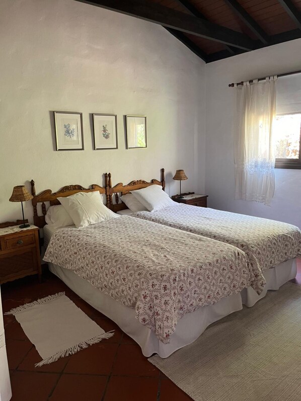 A bedroom with two beds, a nightstand with a lamp, and a window with curtains. - Casa Del Lomo (San Sebastián de La Gomera)