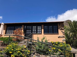 A building with a staircase leading to a door, surrounded by yellow flowers and a clear blue sky. - Casa Del Lomo (San Sebastián de La Gomera)