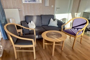 A living room with a grey sofa, two wooden chairs, a round wooden table, and a patterned rug. - Fjord house by the lake (Otterndorf)