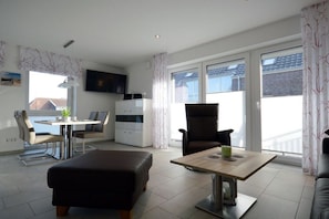 A modern living room with a sofa, a dining table, and a TV mounted on the wall. - Holiday apartment with 1 bedroom (Norddeich)
