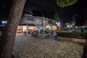 Un patio al aire libre con piso de piedra, mesas y sillas, rodeado de edificios con ventanas iluminadas.