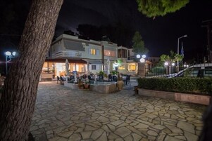 An outdoor patio area with stone flooring, tables, and chairs, surrounded by buildings with illuminated windows.