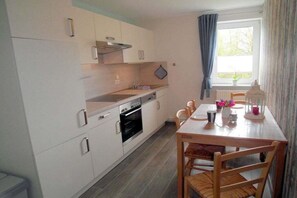 A compact kitchen with white cabinets, a built-in oven, and a dining area with a wooden table and chairs. - Holiday apartment with 2 bedrooms (Vollerwiek)