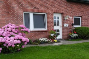 Ein Ziegelhaus mit weißer Tür, zwei Fenstern und einem kleinen Garten mit rosa Hortensien und verschiedenen anderen Blumen.