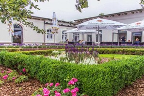 An outdoor seating area with umbrellas, a building with a white facade, and a well-maintained garden with hedges and flowers. - Holiday apartment with 2 bedrooms (Norderney)