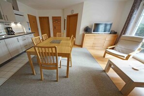 A modern kitchen with white cabinets, a dining table with chairs, a television on a stand, and a window with curtains. - Holiday apartment with 2 bedrooms (Prerow)