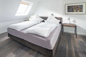 Ein Schlafzimmer mit Bett, Nachttisch, Lampe, einem Gemälde an der Wand und einem Oberlicht.