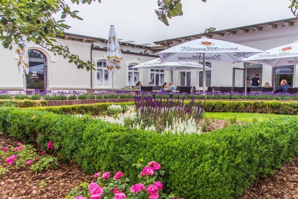 An outdoor seating area with umbrellas, a building with a white facade, and a well-maintained garden with hedges and flowers.