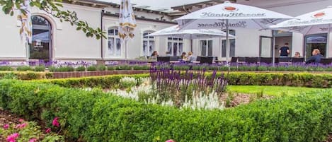 Un área de estar al aire libre con sombrillas, un edificio de fachada blanca y un jardín bien cuidado con setos y flores.