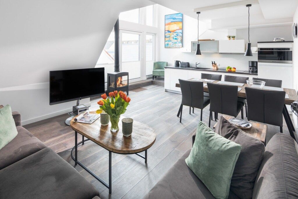 A modern living room with a flat-screen TV, a wooden coffee table with a vase of flowers, and a sofa with cushions.