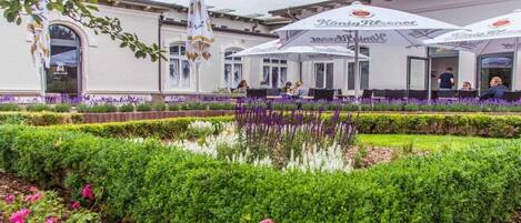Un área de estar al aire libre con sombrillas, un edificio de fachada blanca y un jardín bien cuidado con setos y flores.