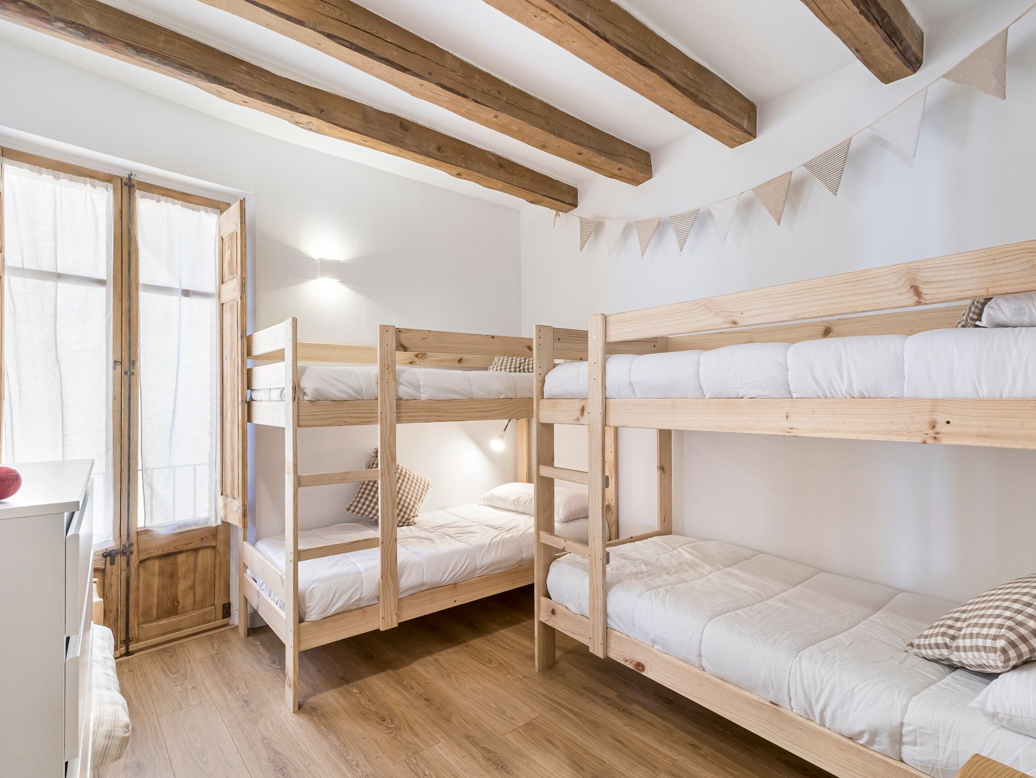 A bunk bed room with wooden beams, white walls, and a window with curtains.