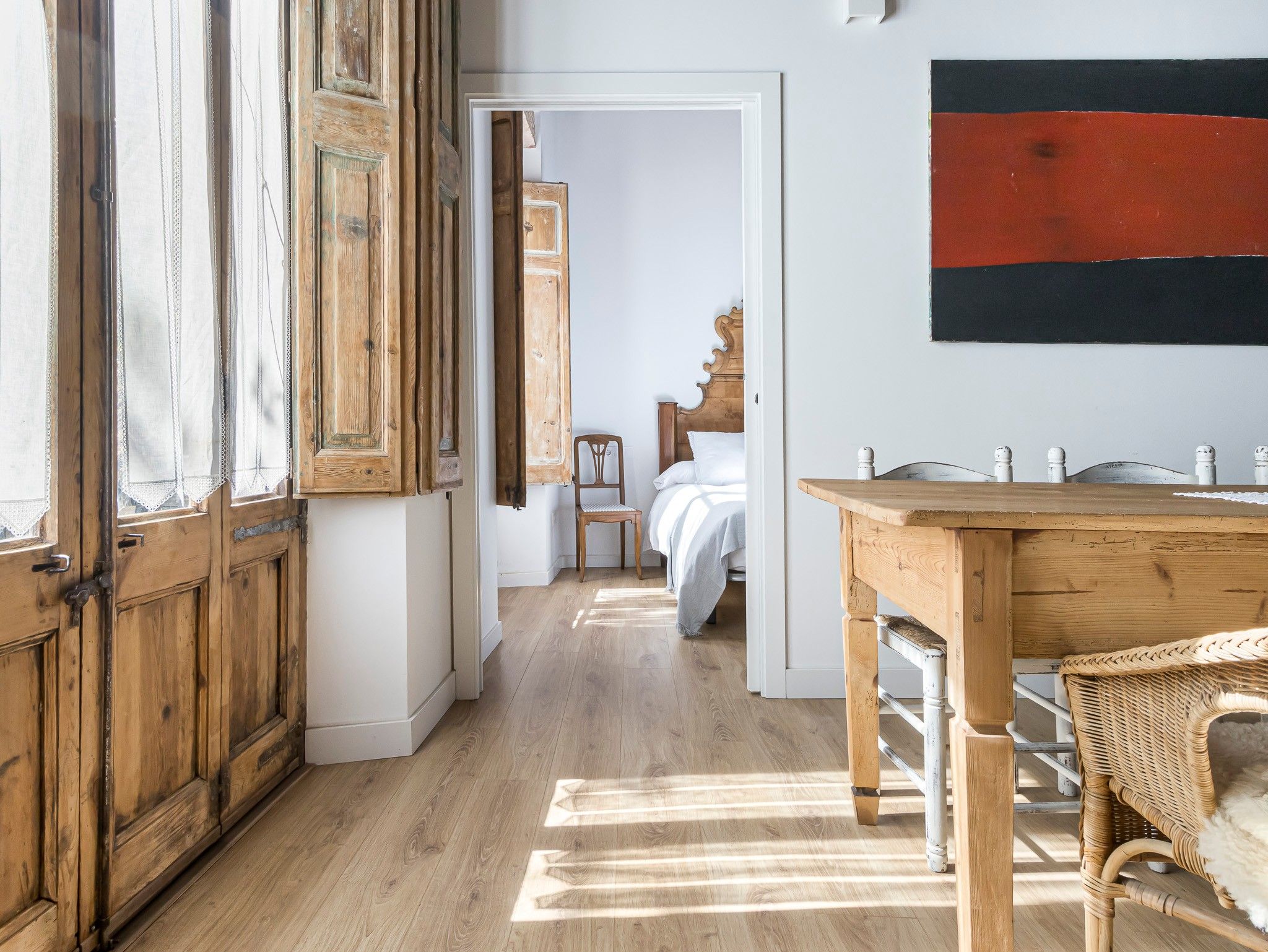 A room with a wooden floor, a bed with white bedding, a wooden table, and a wicker chair.
