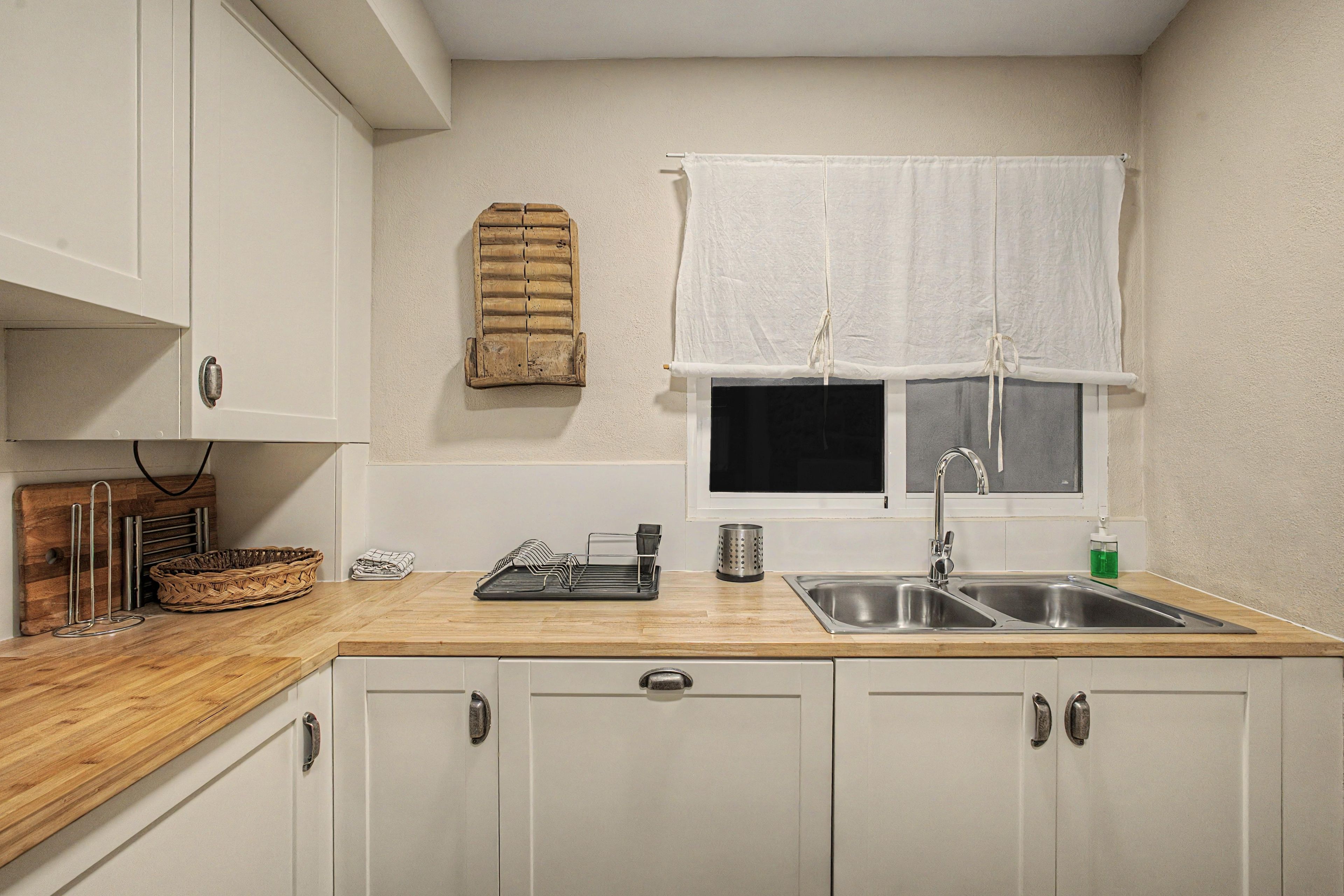 A kitchen with white cabinets, a wooden countertop, a sink, a window with a white curtain, and a hanging wooden object.