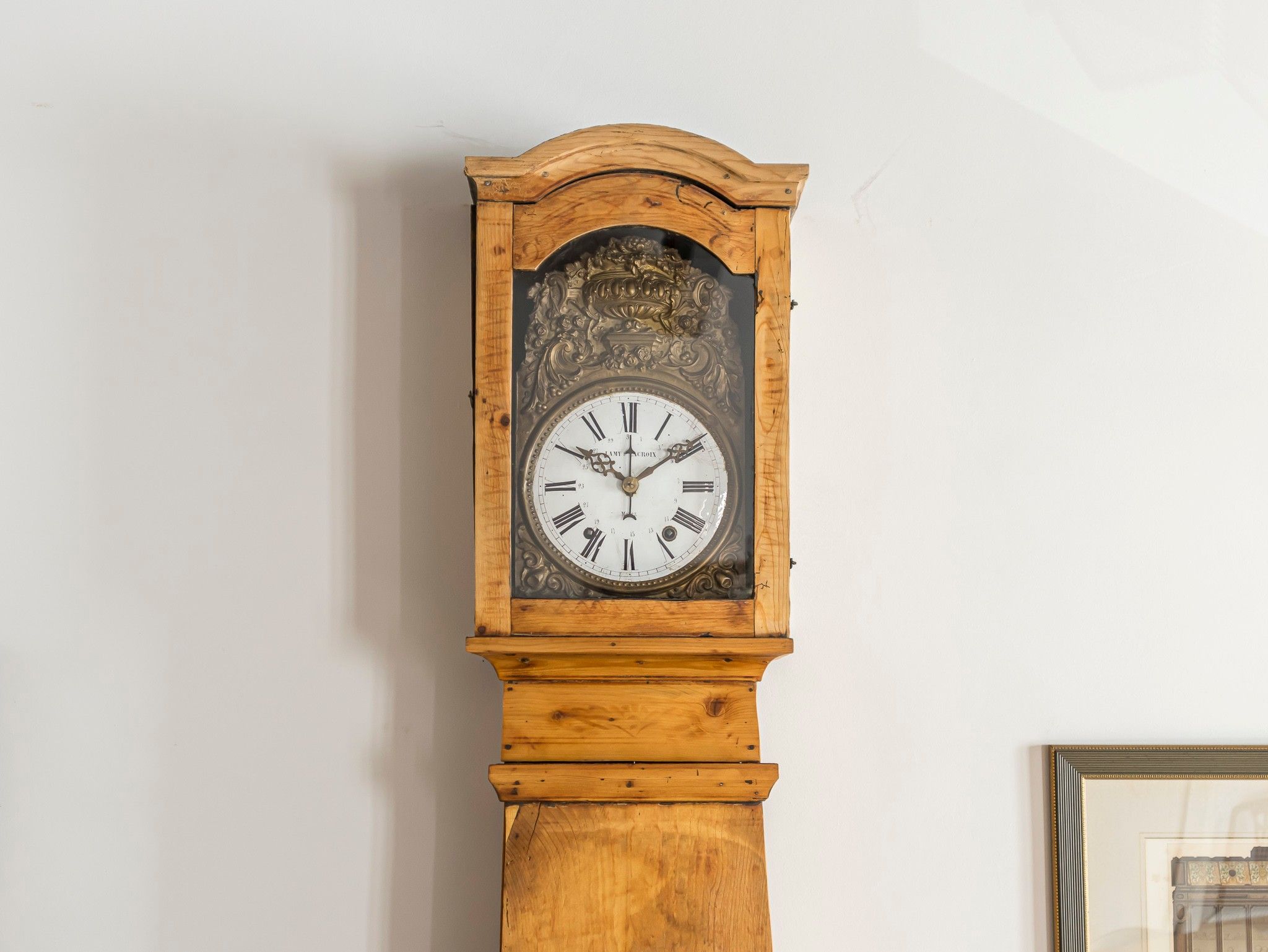 A tall grandfather clock with a wooden base and ornate carvings, mounted on a plain wall.
