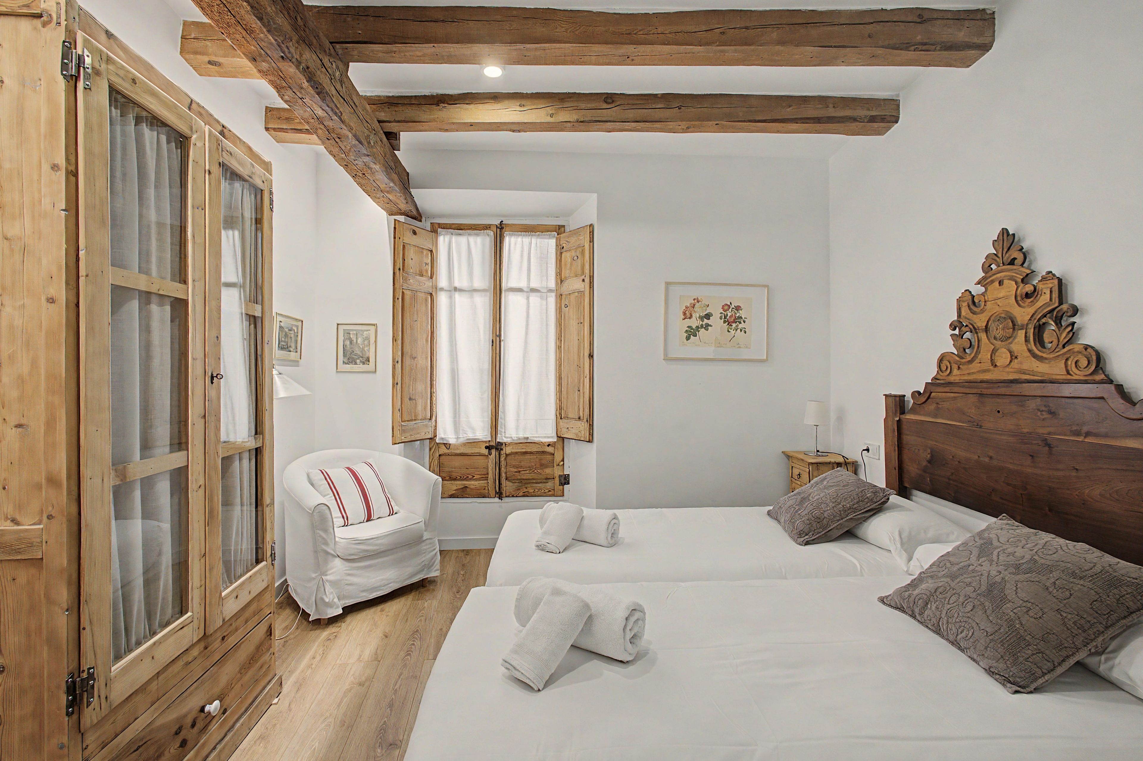 Een slaapkamer met een houten plafond, een bed met witte lakens, een houten hoofdbord, een stoel en een raam met gordijnen.