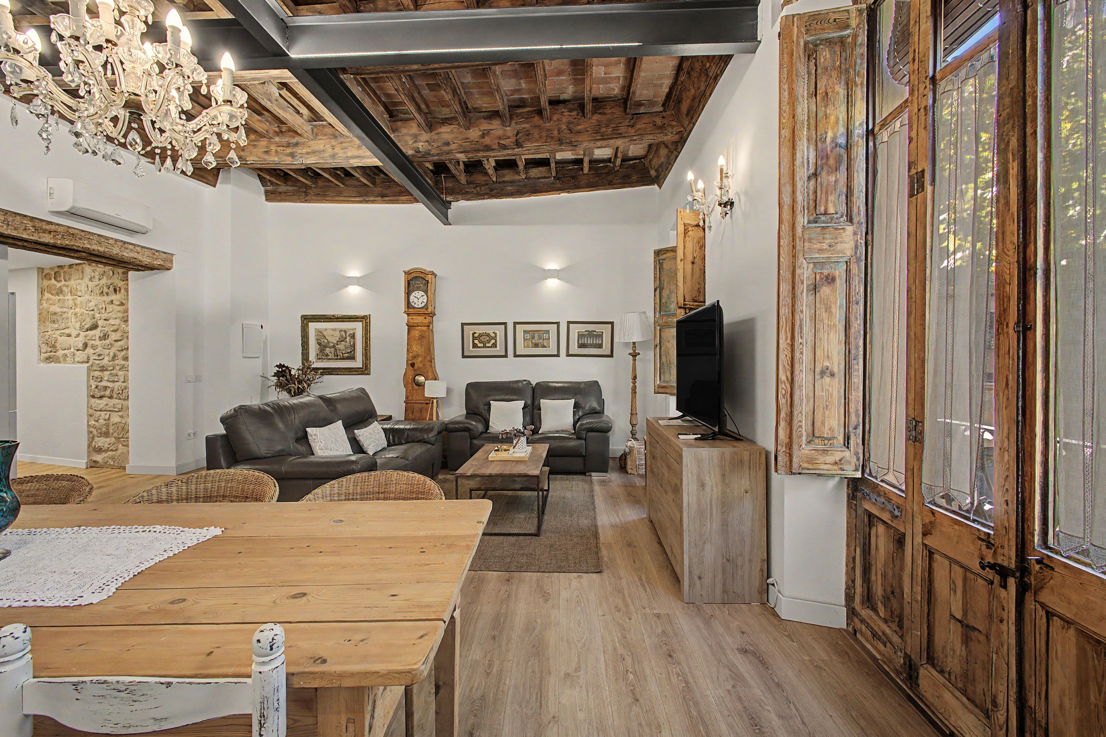 A rustic living room with exposed wooden beams, a stone wall, a wooden dining table, leather sofas, and a television.