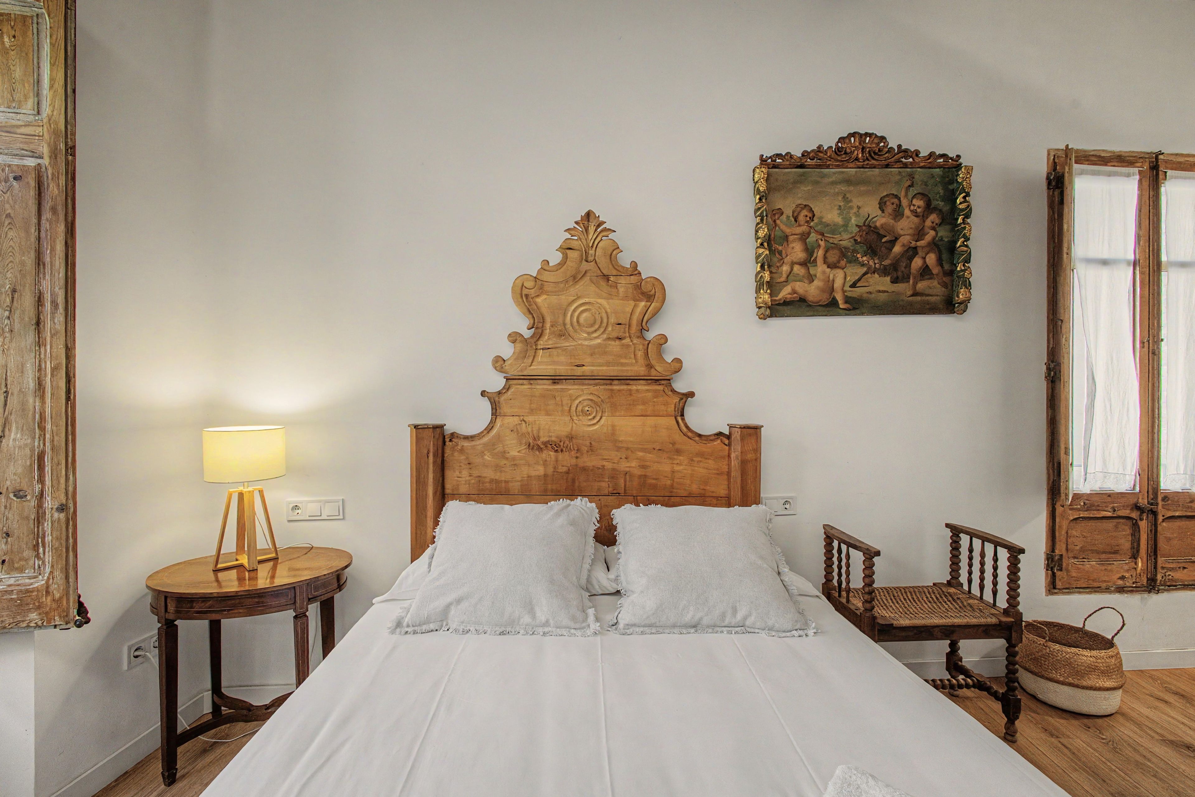 A bedroom with a wooden headboard, a bed with white linens, a round nightstand with a lamp, a wicker chair, and a painting on the wall.