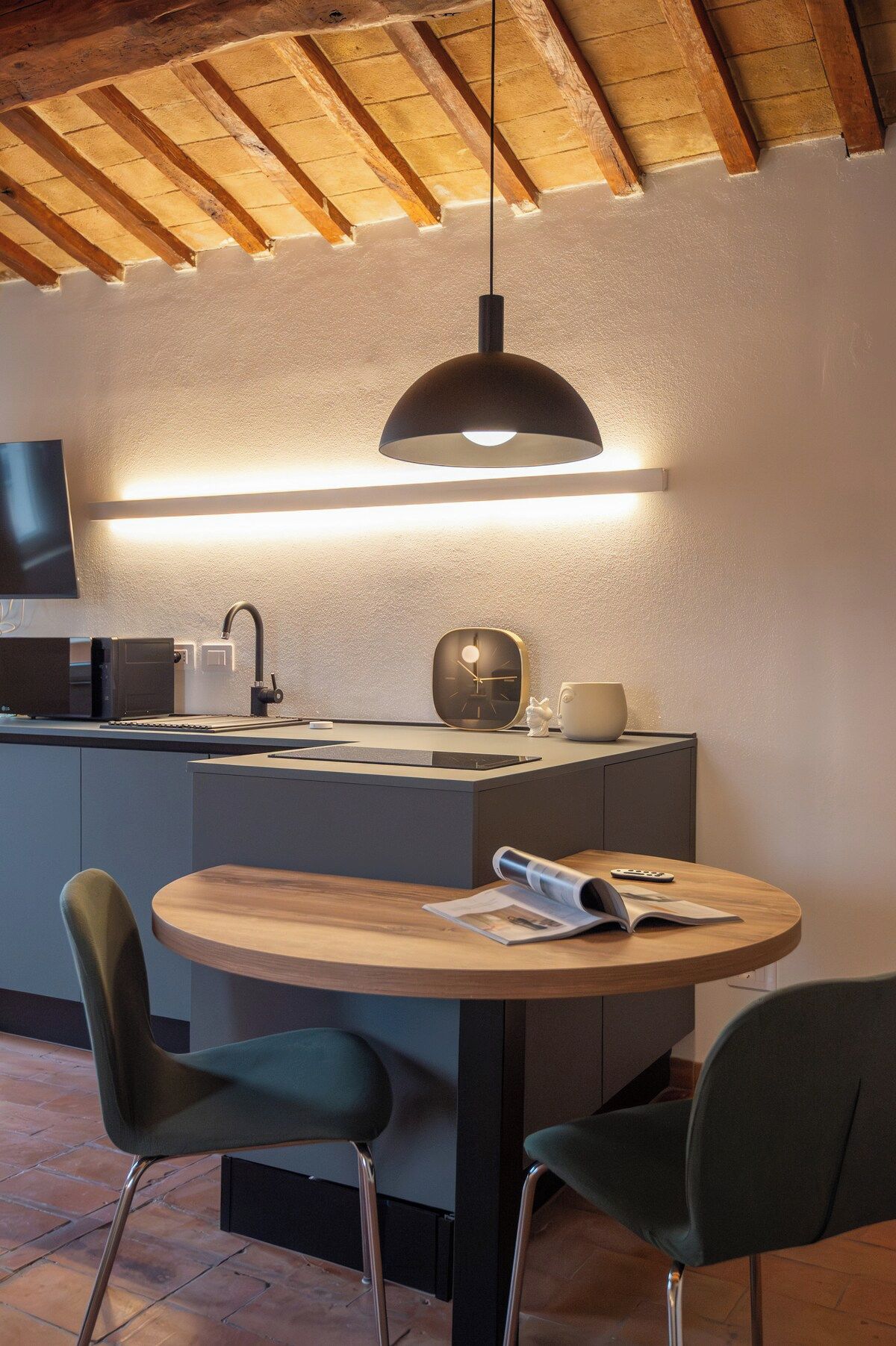 Et moderne køkken med et rundt spisebord, to stole, en hængende lampe, et ur og en kaffemaskine.