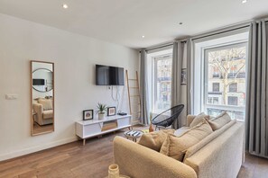 Ein modernes Wohnzimmer mit einer Couch, einem Fernseher, einer weißen Konsole und einem großen Fenster mit Vorhängen.