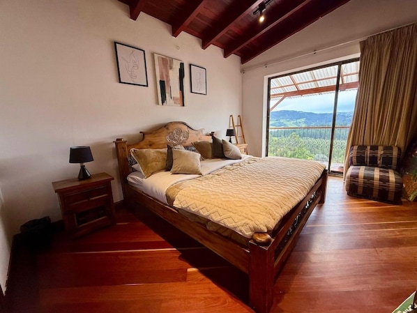 Ein Schlafzimmer mit einem großen Bett, einem Nachttisch, einer Lampe und Blick ins Freie durch ein Fenster.