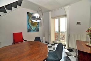 A dining area with a wooden table and chairs, a red cushioned chair, a pendant light, a painting on the wall, and a view through a glass door.