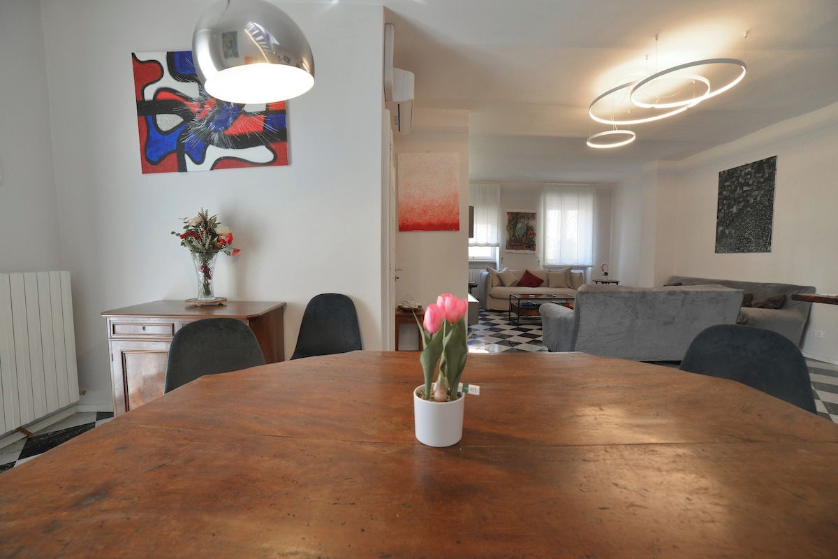A dining area with a wooden table, chairs, and a vase with flowers. There is a painting on the wall and a modern light fixture above the table.