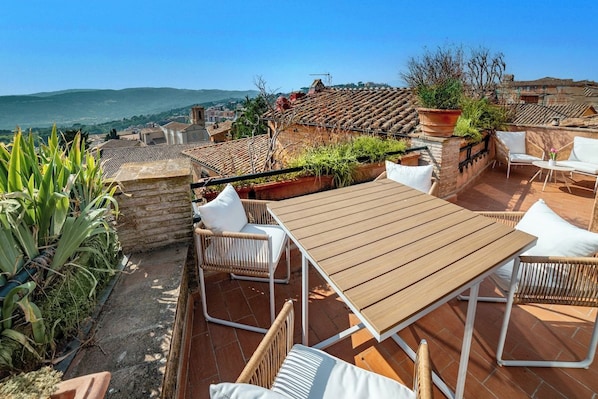 A rooftop terrace with a wooden table and wicker chairs, overlooking a cityscape with terracotta roofs.