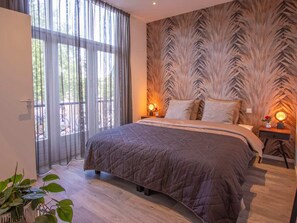 A bedroom with a large bed, a window with curtains, a wall with a leaf pattern, and a bedside table with a lamp. - Do43 "t Groentje 9A (Domburg)