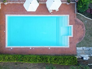 Eine Vogelperspektive zeigt einen rechteckigen Swimmingpool mit Ziegelbordierung, umgeben von einer Hecke und einer Steinmauer.