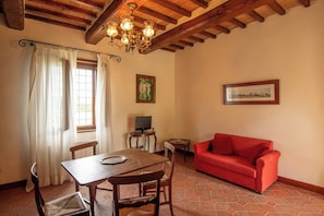Ein Essbereich mit Tisch und Stühlen, ein rotes Sofa, ein Fernseher auf einem Beistelltisch und ein Fenster mit Vorhängen.