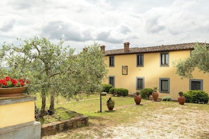 A yellow building with a tiled roof, surrounded by a garden with various plants and a large flower pot. - Villa Missiano Unità Pegaso con Piscina (Panicale)