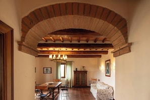 A dining area with a brick arch, wooden beams, a chandelier, a dining table with chairs, a cabinet, and a sofa. - La Fenice Apartment: Pool, BBQ, Free Parking (Panicale)