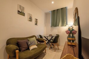 Ein Wohnzimmer mit einem grünen Sofa, einem kleinen runden Tisch und Stühlen. An den Wänden befinden sich Kunstwerke und ein Spiegel.