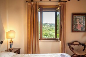 A hotel room with a bed, a nightstand with a lamp, a window with curtains, and a painting on the wall. - Villa Missiano Unità Cassiopea con Piscina (Panicale)